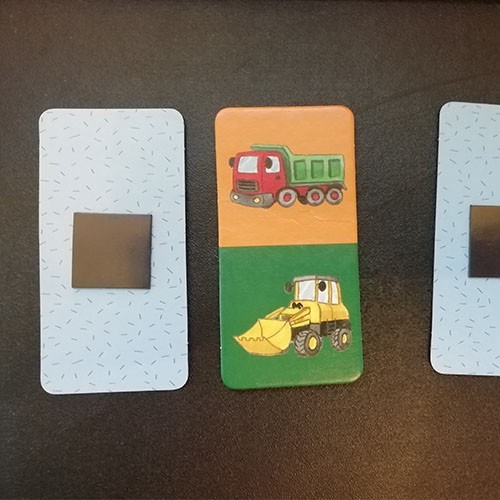 Drei Dominokarten: Zwei sind von der Rückseite zu sehen, auf der Magnete kleben, eine Karte ist von der Motivseite zu sehen, auf der ein LKW und ein Bagger zu sehen sind.