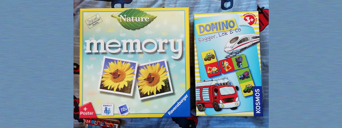 Zwei Kartons von Kinderspielen liegen auf einer Kinderdecke. Das eine ist ein Memory-, das andere ein Domino-Spiel.
