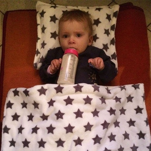 Baby mit Flasche im Mund liegt auf einer Decke und ist zugedeckt mit einer Sternchenbettwäsche.