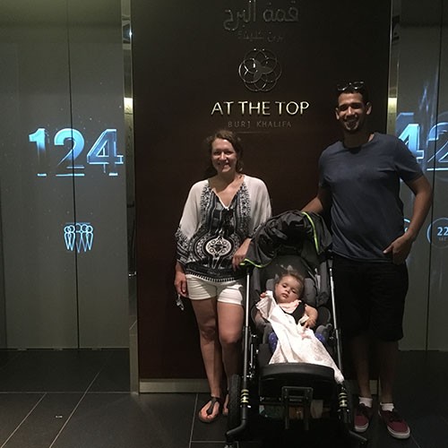 Vater und Mutter stehen im Urlaub in Dubai in einem Gebäude vor den Aufzügen. Vor ihnen steht der Regabuggy, in dem ihre Tochter sitzt und hinter ihnen steht „AT THE TOP, BURJ KHALIFA“ an der Wand.