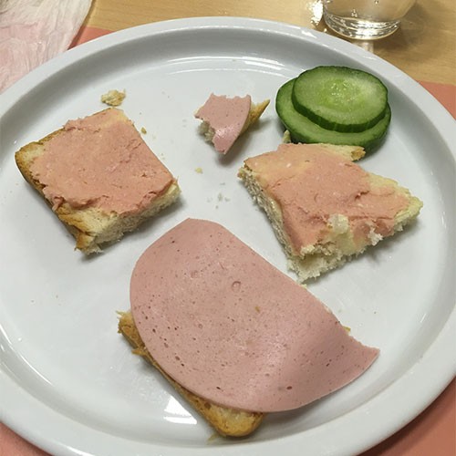 Auf einem Teller liegen Gurkenscheiben sowie große und kleine Stücke Brot, die belegt sind mit Fleischwurst und einer Streichwurst.