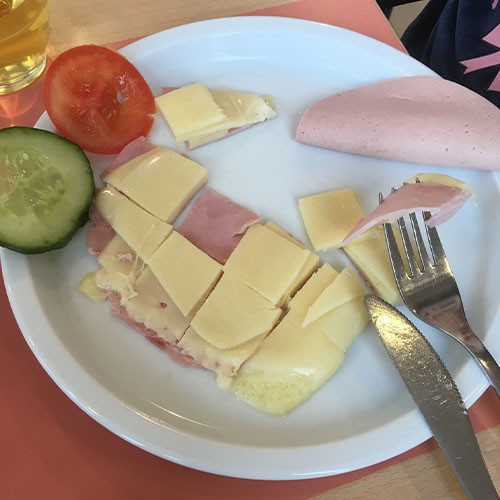 In kleine Häppchen geschnittene Wurst- und Käse-Scheiben, eine Tomaten- und eine Gurkenscheibe sowie Besteck liegen auf einem Teller.