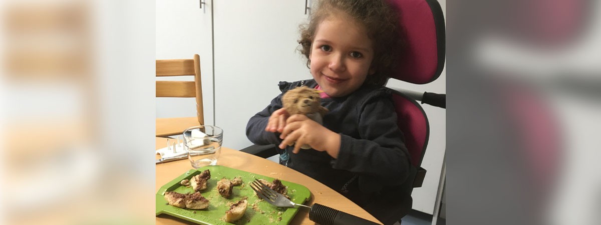 Kleine SMA-Patientin ist in der Reha und sitzt am Tisch. Vor ihr ist ein grünes Brett mit Brotstückchen und einer therapeutischen Gabel. Sie lächelt und hält einen Teddy in den Händen.