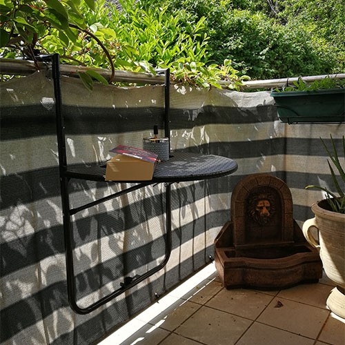 Ein Balkon mit gestreiftem Sichtschutz. An dem Geländer hängt ein halbrunder Tisch, auf dem ein Buch liegt. In der Ecke steht ein kleiner Brunnen mit Löwenkopf. Im Hintergrund sind Büsche und Baumkronen.