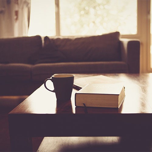 Auf einem Couchtisch im Wohnzimmer befinden sich Bücher und eine Kaffeetasse. Dahinter ist ein Sofa zu sehen. Die Sonne scheint durch die hellen Fenster und die Balkontür.