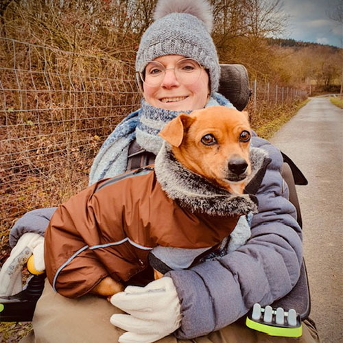 Eine SMA-Patientin sitzt im Rollstuhl und hat ihren Hund auf dem Schoß sitzen. Sie und ihr Haustier tragen warme Kleidung, denn es ist Winter und kalt draußen.