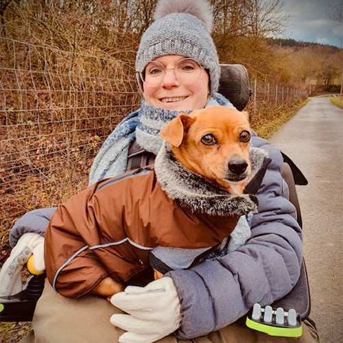 Eine SMA-Patientin sitzt im Rollstuhl und hat ihren Hund auf dem Schoß sitzen. Sie und ihr Haustier tragen warme Kleidung, denn es ist Winter und kalt draußen.