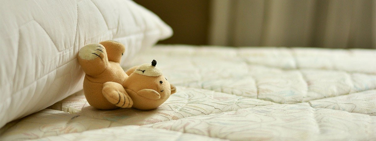 Ein kleiner Teddy-Bär liegt auf dem Rücken im Bett. Matratze und Kissen sind nicht bezogen.