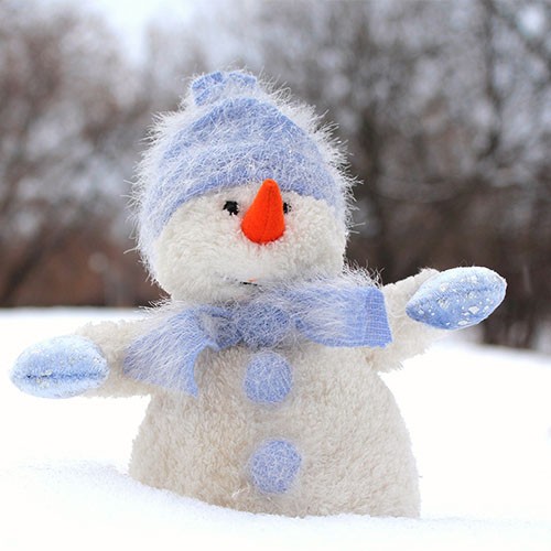 Ein Schneemann aus Stoff steht im Winter im Schnee und hat eine blaue Pudelmütze, Handschuhe und einen Schal an.