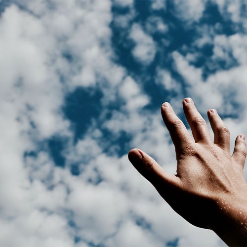 Eine Hand greift in die Luft als wolle sie nach Träumen greifen wollen. Im blauen Himmel ziehen weiße Wolken.