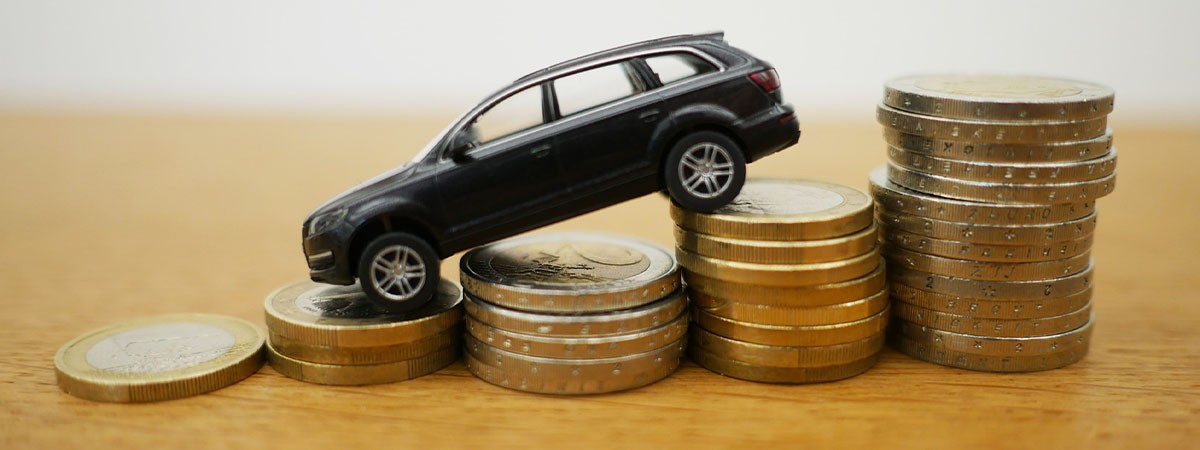 Ein schwarzes Spielzeug-Auto steht auf Stapeln aus Euro-Münzen. Das Vorderteil des Autos steht auf einem niedrigeren Stapel. Dadurch neigt sich das Auto nach unten.