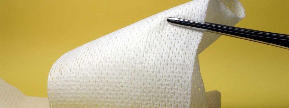 Eine Mullbinde für den Verband von Wunden wird von einer Pinzette vor einem gelben Hintergrund gehalten.
