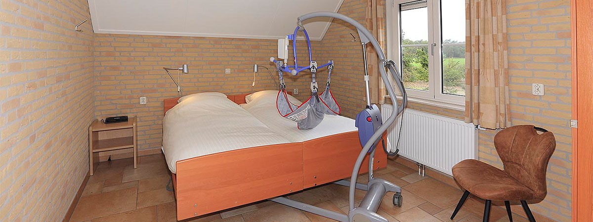 Ein barrierefreies Schlafzimmer in einer holländischen Ferienunterkunft. Vor dem behindertengerechten Doppelbett steht ein Gerät zum Anheben von Patienten.