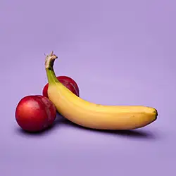 Auf einem violetten Hintergrund liegt eine Banane an zwei Pflaumen angelehnt. 