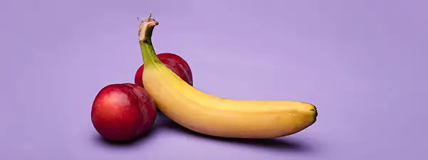 Auf einem violetten Hintergrund liegt eine Banane an zwei Pflaumen angelehnt. 