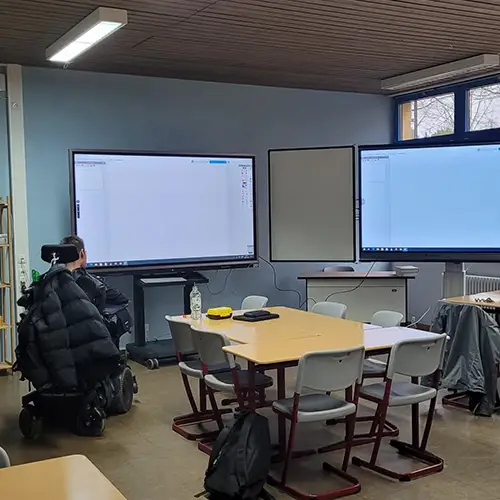 Foto eines Klassenzimmers, in dem mehrere Tischgruppen mit Stühlen stehen. Vor der Wand stehen große digitale Tafeln oder Bildschirme. Links im Bild befindet sich  Autor in seinem Rollstuhl mit dem Rücken zur Kamera und Blick auf die Bildschirme.   