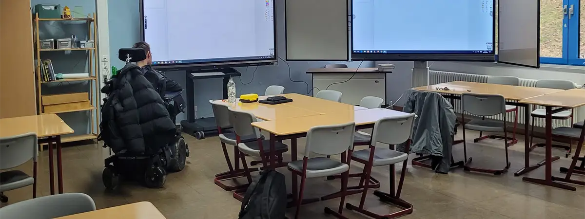Foto eines Klassenzimmers, in dem mehrere Tischgruppen mit Stühlen stehen. Vor der Wand stehen große digitale Tafeln oder Bildschirme. Links im Bild befindet sich  Autor in seinem Rollstuhl mit dem Rücken zur Kamera und Blick auf die Bildschirme.   
