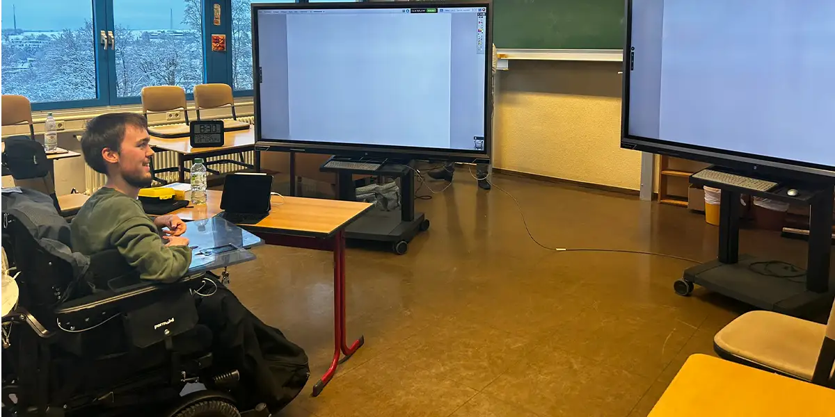 Foto in ein Klassenzimmer, in dem ein paar Schultische und Stühle zu sehen sind, sowie zwei große Bildschirme. Der Autor sitzt im Rollstuhl mit Blickrichtung zu den beiden großen Bildschirmen. 